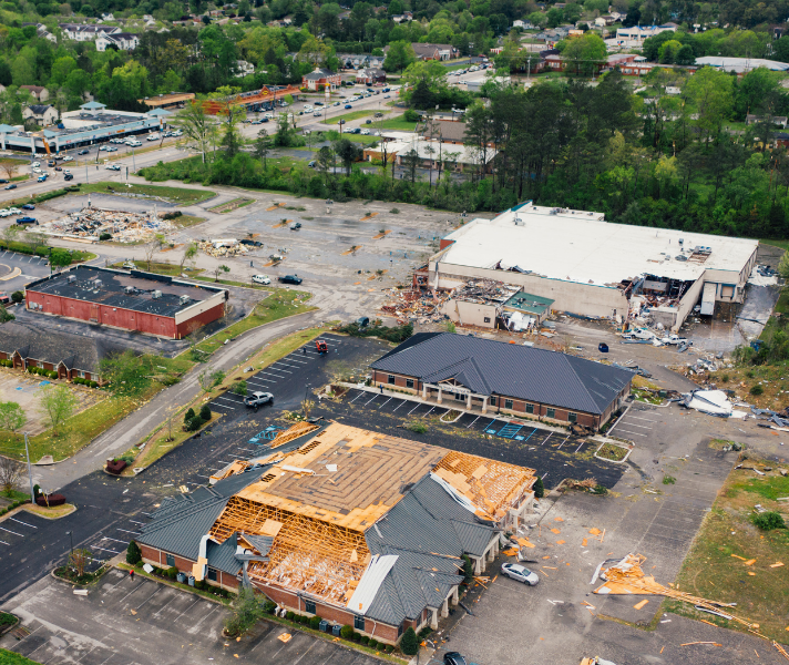 Aerial view of a commercial area with extensive roof damage and scattered debris after a severe storm or disaster, cars parked around damaged buildings